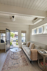 A bright traditional cottage living room with a soft gray sofa, cozy throw, and round marble coffee table sits by an open door. Natural light flows in through windows.