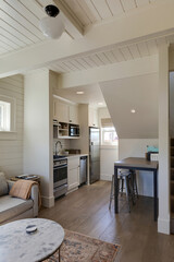 This traditional cottage interior features a compact, efficient kitchen with white cabinetry, stainless steel appliances, and a cozy dining nook with bar stools under the staircase.