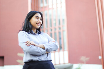 image of successful asian business woman standing over office building with cross hand wearing formal shirt, female entrepreneur smiling confident looking away show optimistic expression