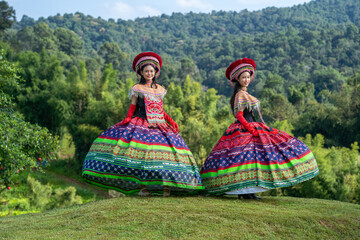 Two women wearing vibrant traditional dresses stand outdoors on a grassy hill, surrounded by lush...