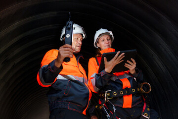 Male and female engineers with reflective orange safety vests working together in large pipes,...