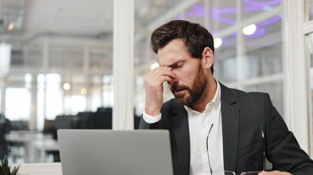 Tired businessman takes off his glasses and rubs his eyes while working on his laptop in the office. He takes a moment to relax and relieve his eye strain from staring at a computer screen.
