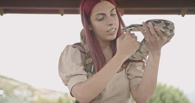 A young woman with red hair smiles as she holds her pet snake. She gently pets the reptile as it wraps itself around her shoulders.