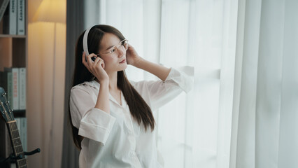 Fototapeta premium Asian woman wearing headphones and glasses is sitting in front of a window. She is listening to music and she is enjoying herself