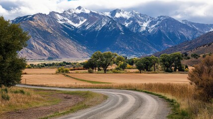 Fototapeta premium A mountain landscape with a winding dirt road leading through fields and forests, framed by majestic peaks.