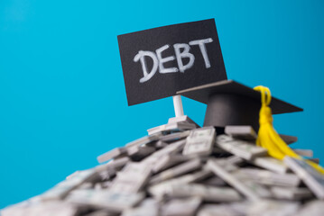 Mortarboard on heap US dollar banknotes and DEBT label with blue background. Student debt or loan,...