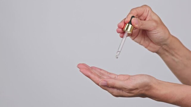 Slow motion close-up of female hands on isolated gray background holding dropper, anti-aging facial serum, booster dripping on palm. Skin care, moisturizing, vitamin c, anti-wrinkle, face lifting ads