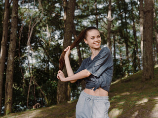 Smiling young woman in casual outfit, playing with her hair in a sunny forest setting Nature and freedom concept, enjoying outdoor lifestyle and tranquility