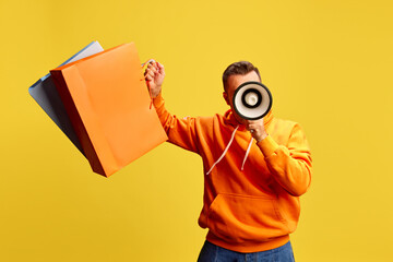 Man enthusiastically holds oversized shopping bags while shouting into megaphone against yellow...