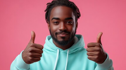 A cheerful man in a mint green hoodie gives a thumbs-up gesture against a pink background, conveying positivity, approval, and a friendly, welcoming demeanor.