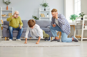 Little boy doing sport exercises lying on floor in living room at home with father and grandfather counting how many push-ups the child has done. Family sport and healthy lifestyle concept.