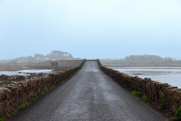 A narrow stone bridge in Connemara, Ireland, extending into the misty distance under an overcast sky, with wet road surface reflecting the foggy ambiance of a quiet, rural landscape