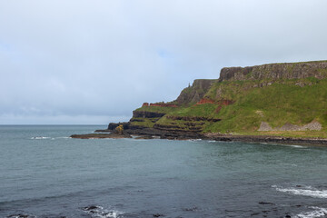 The rugged coastline of Giant Causeway, Northern Ireland, highlights green cliffs and rocky formations, with the Atlantic Ocean extending into the horizon under cloudy skies - Powered by Adobe