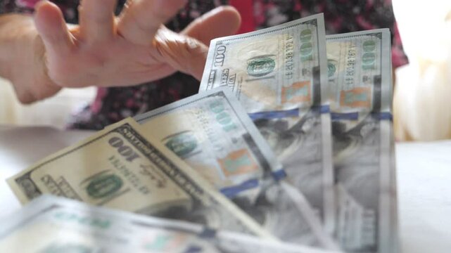 Grandmother holding bundle of money in hands and throwing one hundred dollar bills on the table. Arms of old woman scattering foreign currency on the desk in front to camera. Close up Slow motion