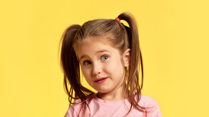 With slightly tilted head and amused expression, girl gives curious look, full of innocence and wonder against vibrant yellow background. Concept of facial-expressions, positive and negative emotions