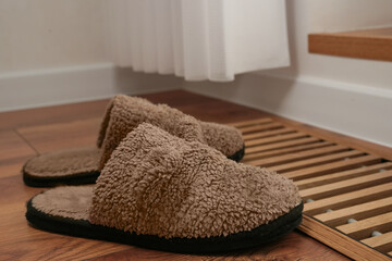 Soft focus of fluffy slippers near protective radiator grille built into the floor for heating panoramic windows. Heating grid with ventilation by the floor in hardwood flooring. Side view. Copy space