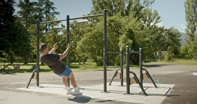 A muscular man performs rows on an outdoor cable machine in a park on a sunny day. He takes off his shirt to reveal a sculpted physique.