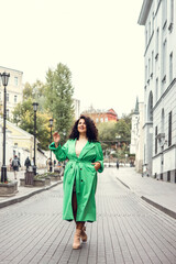 Fototapeta premium Beautiful stylish young woman with curly dark hair posing in a green trench coat in an urban context. Street fashion and style.
