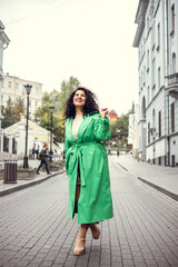 Fototapeta premium Beautiful stylish young woman with curly dark hair posing in a green trench coat in an urban context. Street fashion and style.