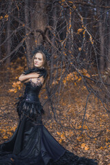 A young woman in the form of a witch and sorceress, standing in a dark forest, wearing a black dress with feathers. A beautiful gothic queen works wonders in the autumn forest on Halloween.