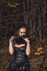 Naklejka premium A young woman in the form of a witch and sorceress, standing in a dark forest, wearing a black dress with feathers. A beautiful gothic queen works wonders in the autumn forest on Halloween.