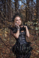 Naklejka premium A young woman in the form of a witch and sorceress, standing in a dark forest, wearing a black dress with feathers. A beautiful gothic queen works wonders in the autumn forest on Halloween.
