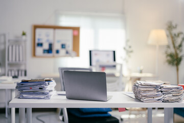 Laptop is sitting on a white desk in a modern office, with piles of paperwork on either side, suggesting a heavy workload and the pressure of approaching deadlines