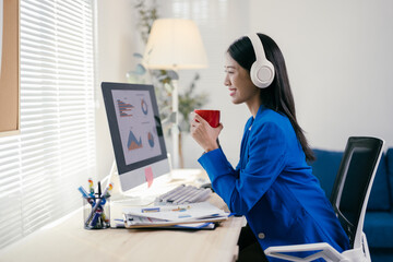 Businesswoman wearing headphones and working from home is taking a coffee break while looking at...