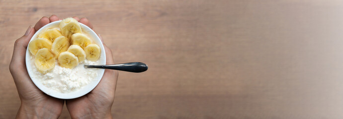 Woman holding a bowl of cottage cheese and banana for a healthy breakfast or snack on a wooden table with copy space. Banner