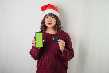 Excited young asian woman wearing santa clause hat and red long sleeved sweater is holding smartphone and credit card, isolated over white background. Concept for Christmas Holiday and New Year Party