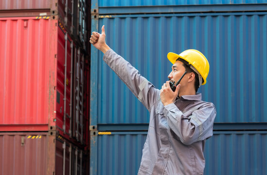 Japanese foreman in yellow hard hat and grey uniform gives thumbs up gesture while speaking on walkie talkie for container loading. Dock worker standing in front of stack of colorful shipping containe