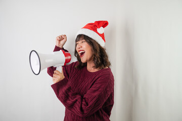 Excited young asian woman wearing santa clause hat and red long sleeved sweater is using megaphone with smiling face expression, isolated on white background. Concept for Christmas and New Year Party