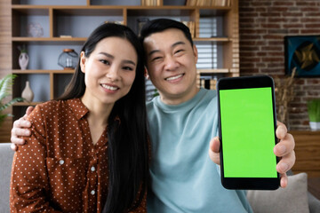 Smiling Asian couple sitting on couch showing green screen smartphone. They appear happy and connected in cozy home setting, highlighting technology, connection, and togetherness.