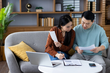 Asian couple reviewing financial documents in living room, illustrating teamwork, home finance...