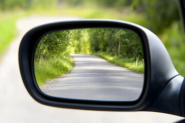 Naklejka premium A close-up of a side mirror reflecting an empty winding road surrounded by lush greenery and trees under bright daylight