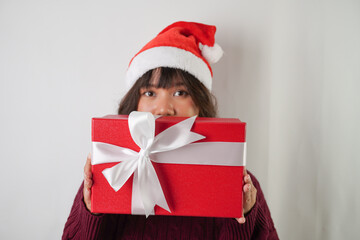Excited young asian woman wearing santa clause hat and red long sleeved sweater is carrying large gift box with ribbon, isolated over white background. Concept for Christmas Holiday and New Year Party