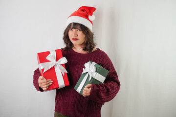 Excited young asian woman wearing santa clause hat and red long sleeved sweater is carrying large gift box with ribbon, isolated over white background. Concept for Christmas Holiday and New Year Party
