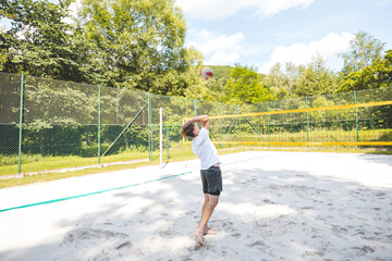 Volleyball player jumping to hit the ball over the net on a sand court during an outdoor game. The action captures the intensity and athleticism of beach volleyball