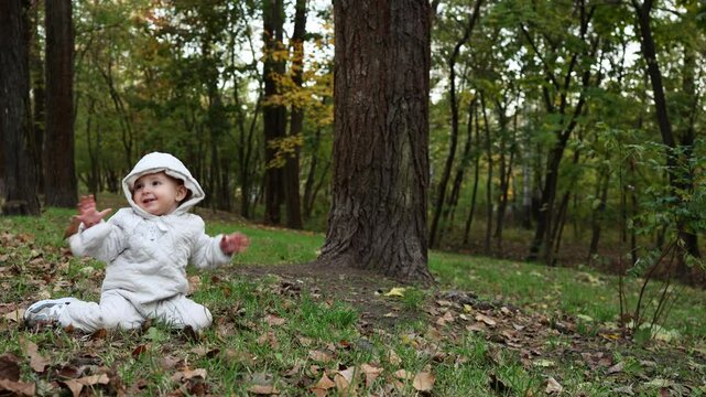 A small baby dressed in a warm white onesie is sitting on the grass in an autumn park, bouncing and joyfully playing.