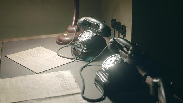 In the semi-darkness there is an old-fashioned Soviet-era table with a lamp, newspapers and black, gloomy rotary telephones. Closeup. Shot in motion