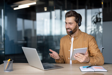 Mature businessman in headset conducts online meeting using laptop and tablet. Professional setup...