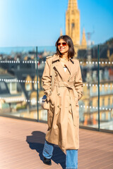 A woman wearing a tan trench coat is walking on a wooden deck