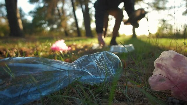 Close-up of plastic litter on grass in a forest with a person cleaning up trash in the background