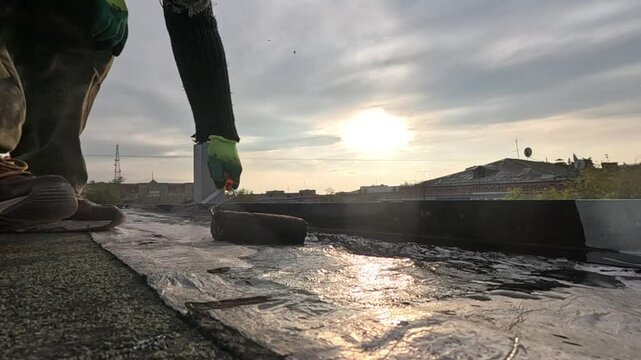 a man priming a roof before repairing the roofing felt, to avoid leaks in the future, against the backdrop of a sunset in an urban development