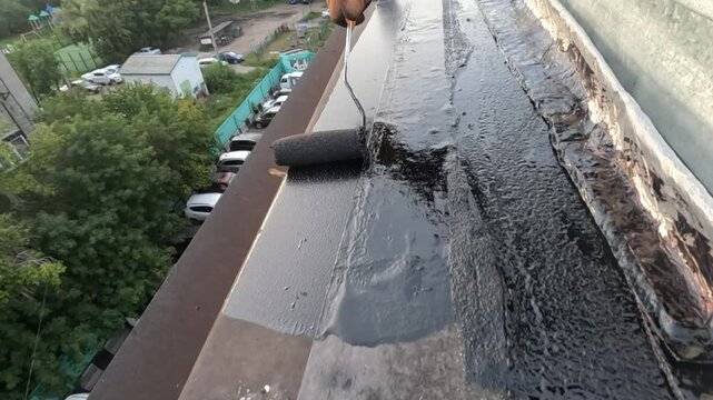 a man priming a roof before repairing the roofing felt, to avoid leaks in the future, against the backdrop of a sunset in an urban development