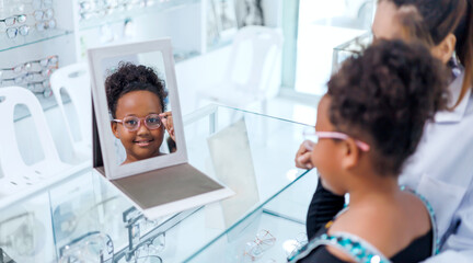 Young african girl getting examine and diagnose by asian female doctor optometrist specialist  choosing fitting correcting eyeglass optical vision for patient in an optician shop.