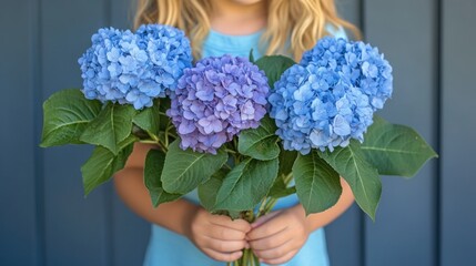 A young girl holds a bouquet of blue and purple hydrangeas in front of a blue wooden wall.