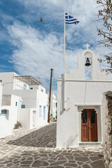 Charming narrow alley leading to a traditional Cycladic church with a Greek flag waving above, located in the picturesque village of Paros, Greece.