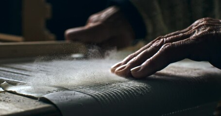 Super slow motion macro of artisan touching threads with flying dust or talc powder particles splashing around, while working with woven weaving machine to craft cloth piece in workshop at 1000 fps. 