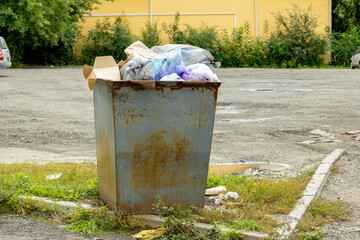 A rusty dumpster overflowing with garbage in a deserted parking lot surrounded by greenery on a sunny day.